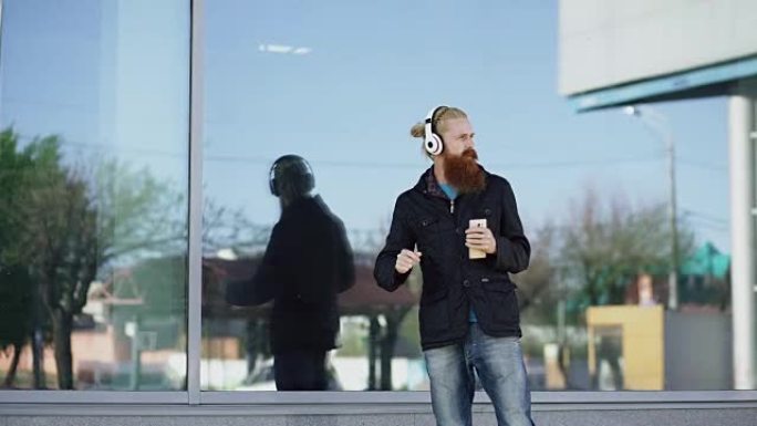 带着耳机和智能手机的快乐年轻大胡子潮人在城市街道上听音乐跳舞