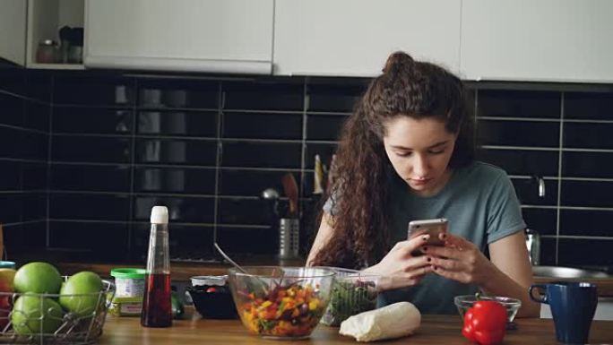 年轻美丽的白人妇女坐在现代轻便宽敞的厨房里的餐桌旁，使用智能手机，发短信，摆放食物的盘子摆在她面前