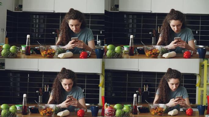 年轻美丽的白人妇女坐在现代轻便宽敞的厨房里的餐桌旁，使用智能手机，发短信，摆放食物的盘子摆在她面前