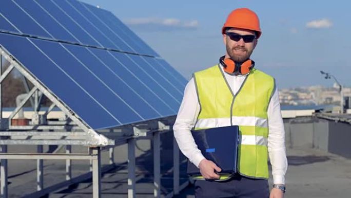 一名工人站在太阳能电池板附近并拿着笔记本电脑时，对着相机微笑。4K。