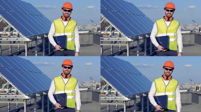 一名工人站在太阳能电池板附近并拿着笔记本电脑时，对着相机微笑。4K。