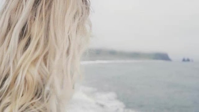年轻的金发女子站在海滩上，靠近冰岛的巨魔脚趾，整理头发，看着水面