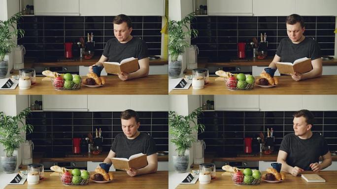年轻的高加索人的肖像坐在现代宽敞厨房的桌子旁，看书，喝咖啡，合上书，透过窗户若有所思地看着