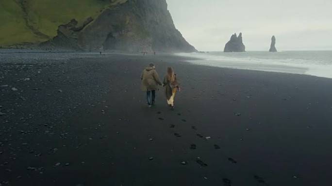 在冰岛巨魔脚趾附近的黑色火山海滩上奔跑的年轻时髦夫妇的鸟瞰图