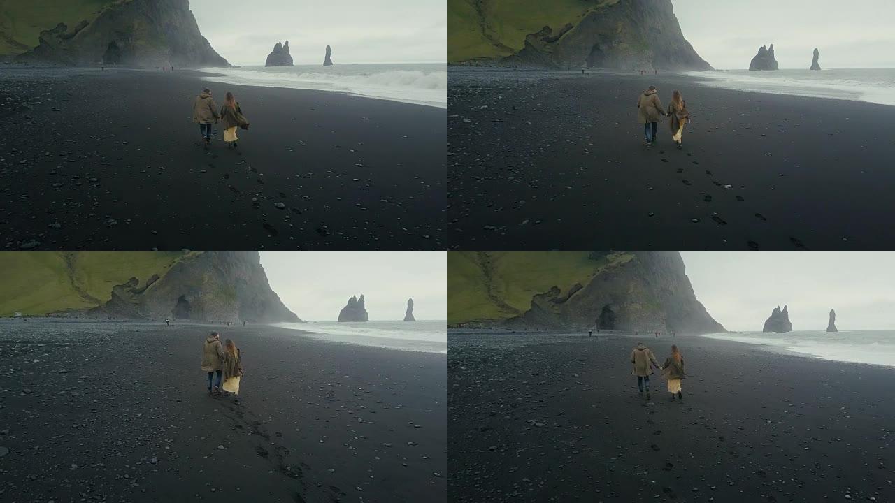 在冰岛巨魔脚趾附近的黑色火山海滩上奔跑的年轻时髦夫妇的鸟瞰图