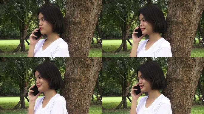 快乐的年轻女子坐在秋公园的树下用手机聊天。