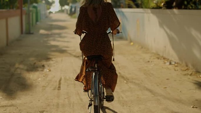 一个穿着连衣裙的美女骑着自行车穿过小镇的狭窄街道。南亚自然，棕榈树生长在路边。慢动作。