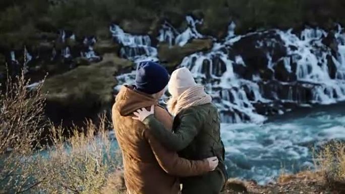 年轻的幸福夫妇看着冰岛的巴纳福斯瀑布，跳起来，举手。男人拥抱和亲吻女人