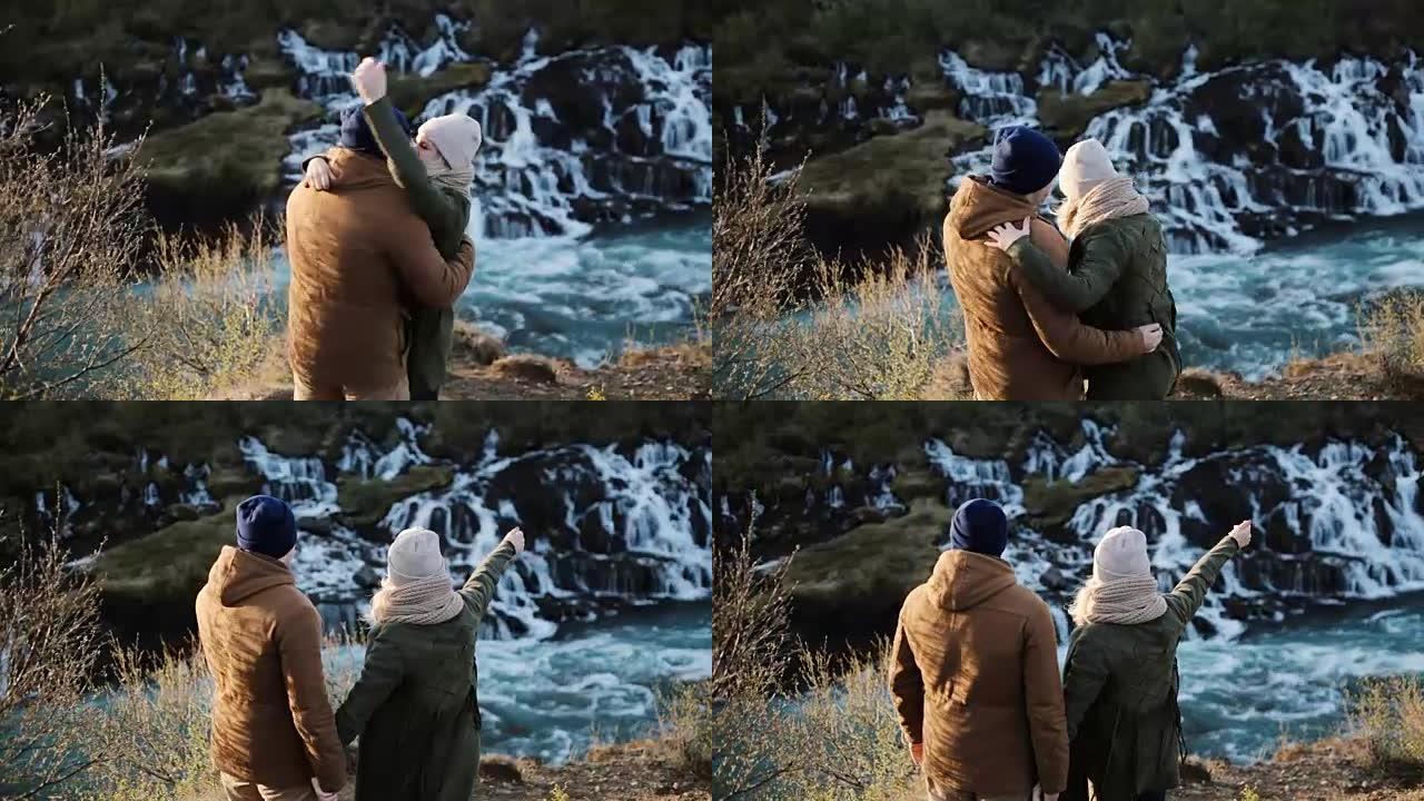 年轻的幸福夫妇看着冰岛的巴纳福斯瀑布，跳起来，举手。男人拥抱和亲吻女人