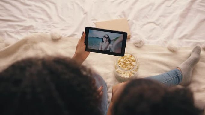 两个女友坐在家里的床上与他们的朋友在海滩度假时在平板电脑上的skype聊天的俯视图