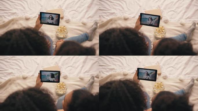 两个女友坐在家里的床上与他们的朋友在海滩度假时在平板电脑上的skype聊天的俯视图