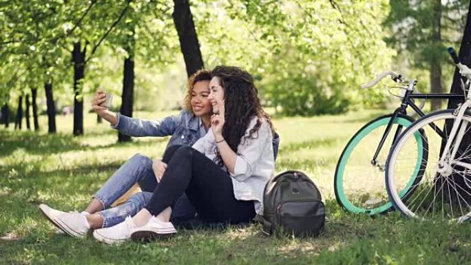 年轻女子和她的非裔美国朋友正在自拍，摆姿势，坐在公园的草坪上玩得开心。温暖的晴天，美丽的大自然和现代