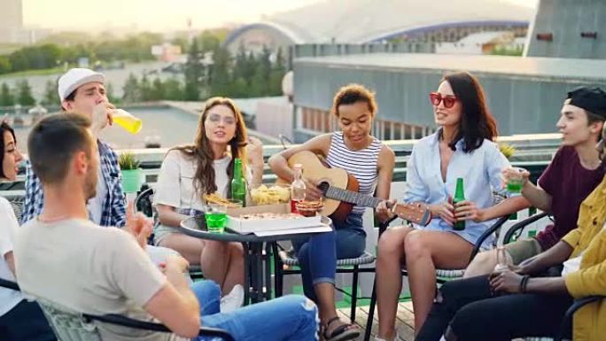 迷人的非洲裔美国女孩正在弹吉他，她的朋友们在屋顶的餐桌上唱歌，喝酒和吃饭，享受周末和温暖的晴天。