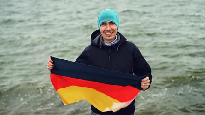 快乐的德国体育迷的慢动作肖像举着德国的国旗，微笑着站在湖边，看着海浪背景下的相机。