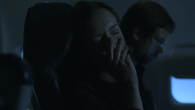 年轻女子正在飞机上睡觉，一名男子坐在窗户旁边的背景中。