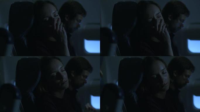 年轻女子正在飞机上睡觉，一名男子坐在窗户旁边的背景中。