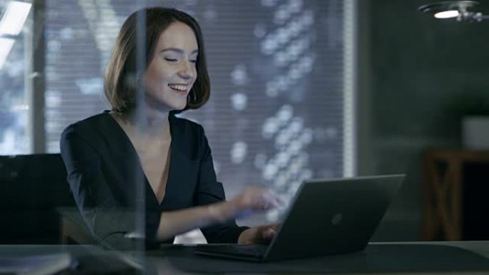 女高管在她的私人办公室里用笔记本电脑工作，可以看到大城市的景色。她笑得很迷人。她的工作空间是用暗淡的