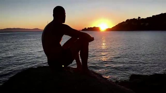 欣赏日落。男人坐在岩石海滩上看着风景
