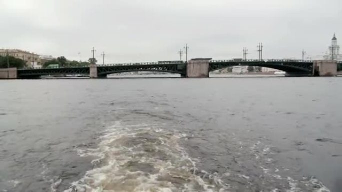 白天的城市景观。从旅途中的一辆河巴士上看到一座桥。水飞溅，天空多云。俄罗斯圣彼得堡