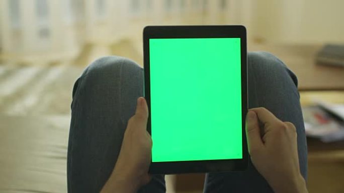 男子躺在家里的沙发上，在膝上的纵向模式下使用带有绿屏的平板电脑