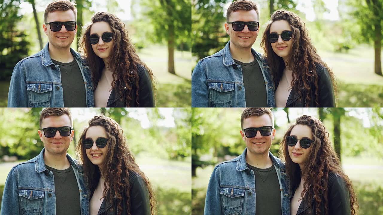 戴着墨镜和时尚服装的幸福夫妇的肖像看着相机，微笑着美丽的绿色公园背景。