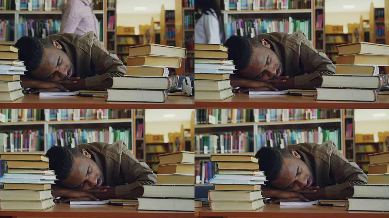 非裔美国英俊的年轻男学生坐在桌子旁，图书馆里堆着一大堆书。他的头躺在桌子上，他睡着了，累了。