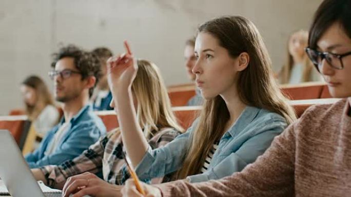 美丽的年轻学生在大学听讲座时使用笔记本电脑，她举手问讲师一个问题。多民族的现代光明学生。