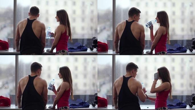 年轻的运动男女站在运动俱乐部的窗户旁聊天。女人在锻炼后喝水