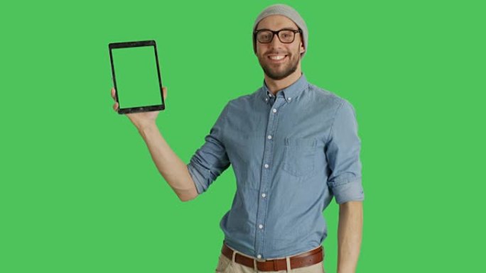 一个戴着帽子和眼镜的时尚男人的中间镜头，一只手拿着平板电脑，另一只手挥动触摸手势。平板电脑和背景是绿