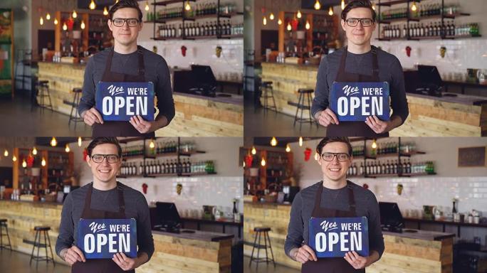 围裙上开朗英俊的年轻服务员的肖像，站在开设咖啡店的 “我们是开放的” 标志上。创业和成功人士的概念。