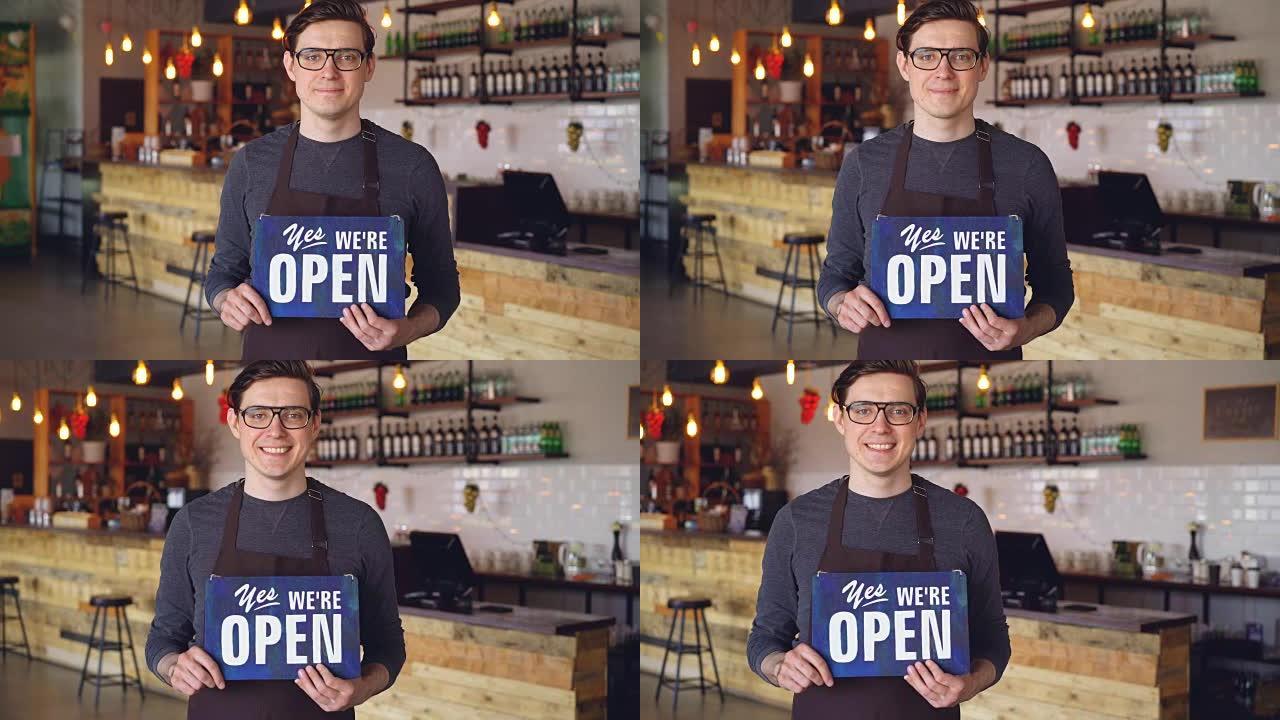 围裙上开朗英俊的年轻服务员的肖像，站在开设咖啡店的 “我们是开放的” 标志上。创业和成功人士的概念。