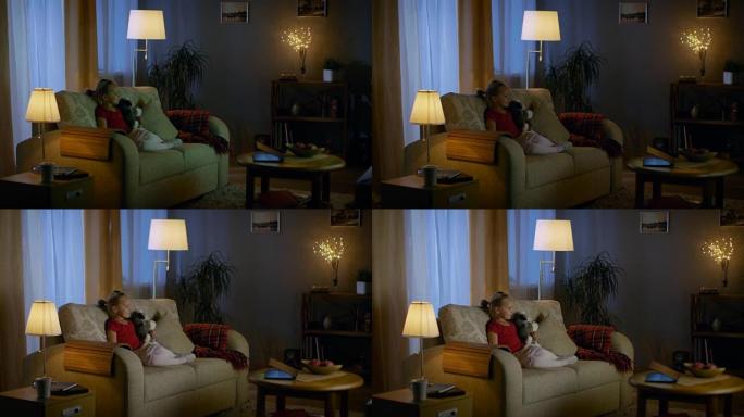 晚上，可爱的小女孩坐在客厅的沙发上，她看电视。她拿着她的毛绒玩具，房间灯亮着。