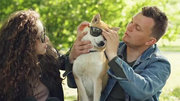 开朗的夫妇在公园里度过周末时，狗戴上墨镜，笑着玩得很开心。自然、现代生活方式和动物概念。