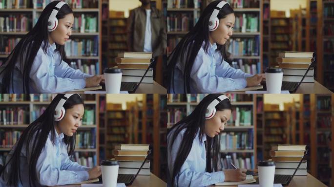 戴着耳机的美丽亚洲女学生坐在图书馆的办公桌上，集中在学校项目中工作。年轻女孩在笔记本电脑上打字，喝咖