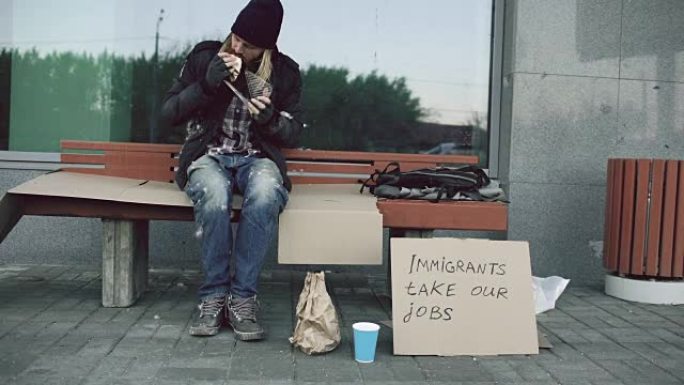 由于欧洲移民危机，无家可归和失业的欧洲男子在城市街道的长凳上吃三明治
