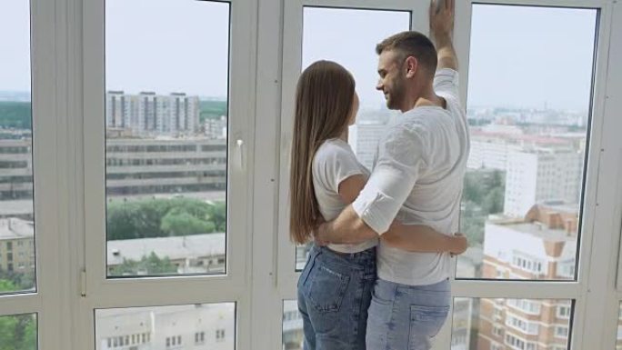 年轻幸福的夫妇拥抱站在窗户附近，欣赏新公寓的景色