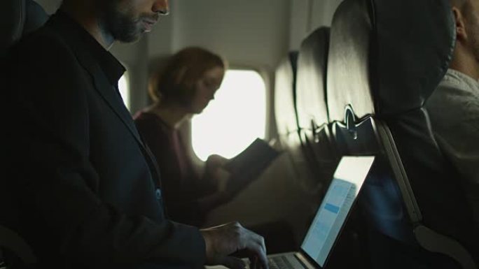 年轻人正在飞机上的笔记本电脑上工作，一个女人正在窗户旁边的背景中阅读。