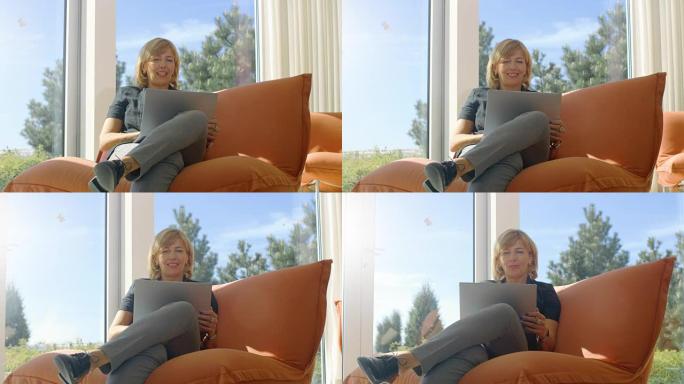 美丽的高级女人坐在阳光明媚的客厅的沙发上使用笔记本电脑。房子有落地窗，可以看到风景。天气晴朗。