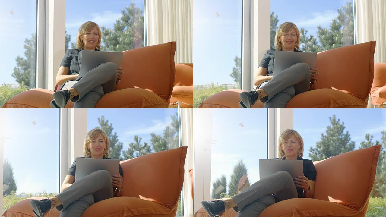 美丽的高级女人坐在阳光明媚的客厅的沙发上使用笔记本电脑。房子有落地窗，可以看到风景。天气晴朗。