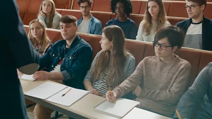 面向镜头的课堂: 大学教授的后视图向充满多民族学生的教室授课。当学生听的时候，讲师用手打手势。