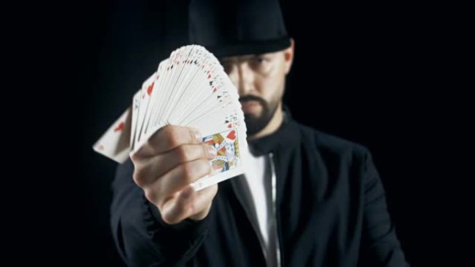 戴着帽子的专业街头魔术师表演手牌技巧，扇出卡片，用手指旋转一张卡片。背景是黑色的。