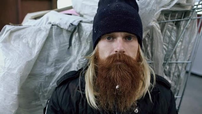 在寒冷的冬日里，年轻的大胡子无家可归的人坐在购物车和垃圾桶附近的人行道上的特写肖像
