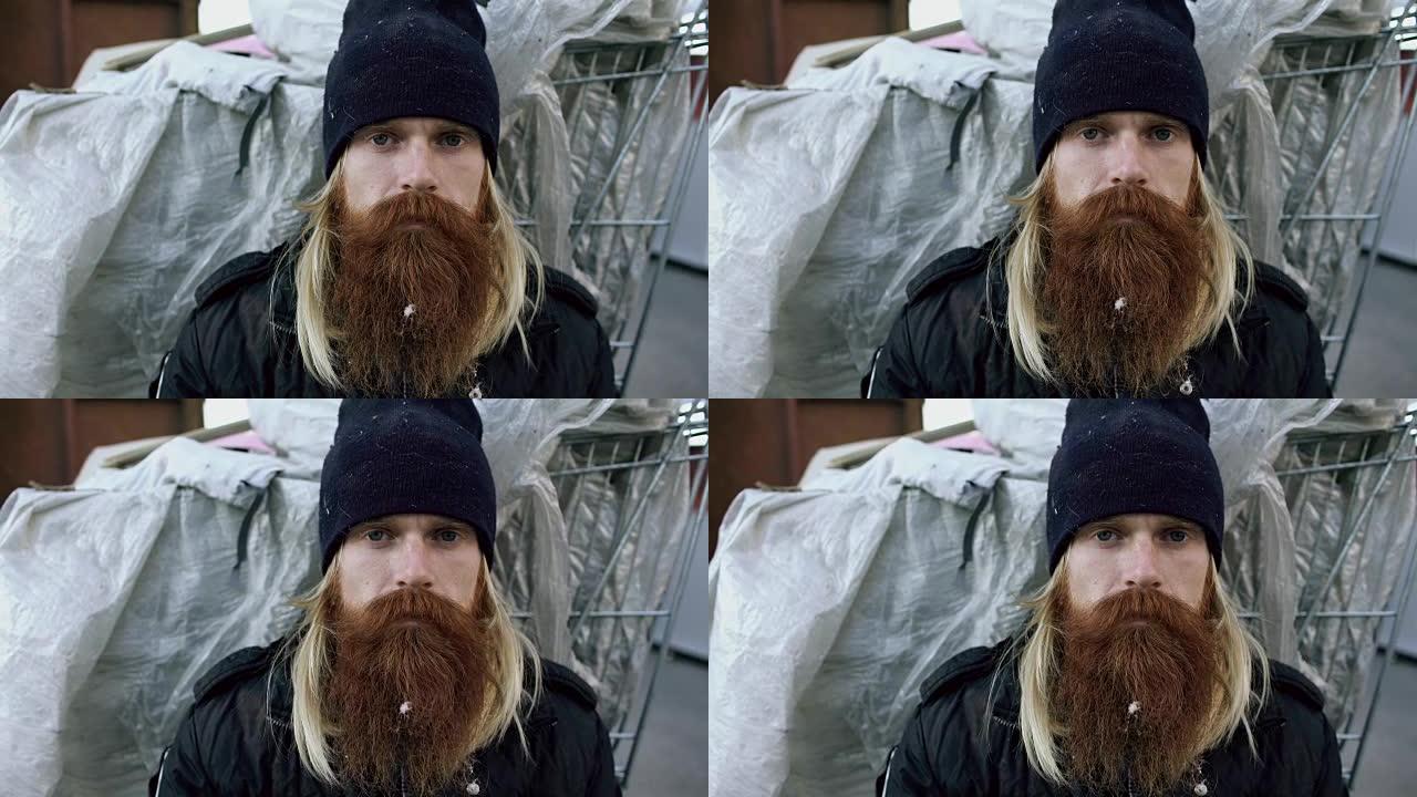 在寒冷的冬日里，年轻的大胡子无家可归的人坐在购物车和垃圾桶附近的人行道上的特写肖像