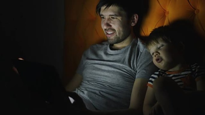 年轻的父亲和他的小儿子用平板电脑看卡通电影，晚上在家躺在床上聊天