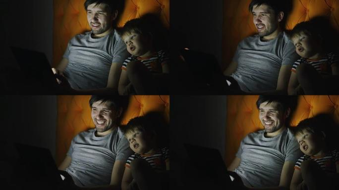 年轻的父亲和他的小儿子用平板电脑看卡通电影，晚上在家躺在床上聊天