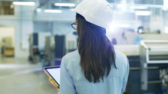 戴着安全帽的漂亮女工业工程师在大工厂散步时使用平板电脑。