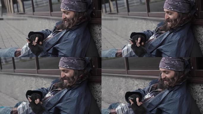 大胡子乞讨的人坐在街上，拿着智能手机