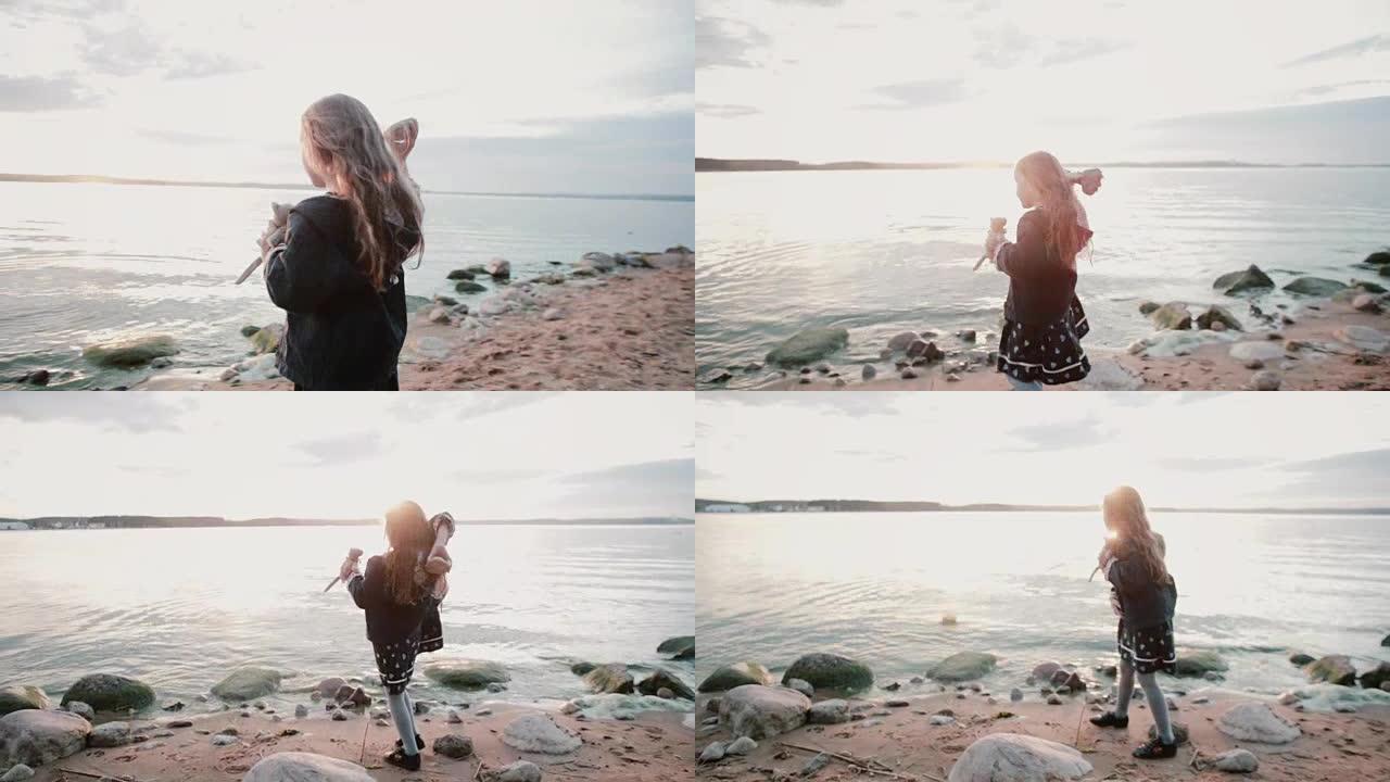 可爱的小女孩站在水边的岸边。女性在日落时向湖中扔一块大石头