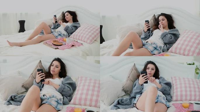 年轻微笑的女人躺在床上。黑发女孩在早餐时上网，使用智能手机和喝果汁