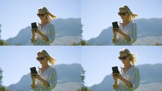 穿着便服和太阳镜的美丽年轻金发女子站在草地上使用手机，背景是丘陵和山脉。现在是夏天。